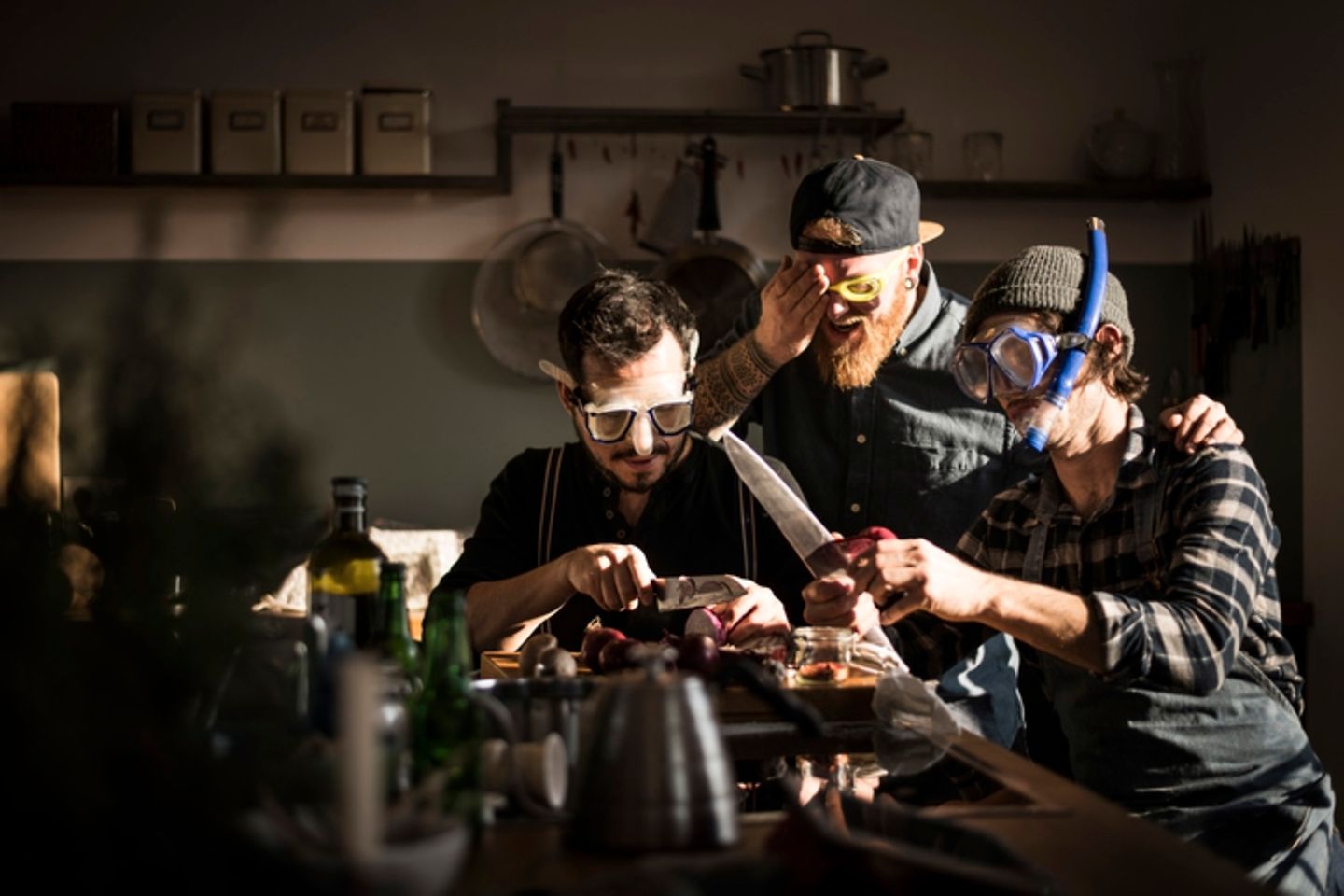 Three men cook together and cut onions with diving goggles on.