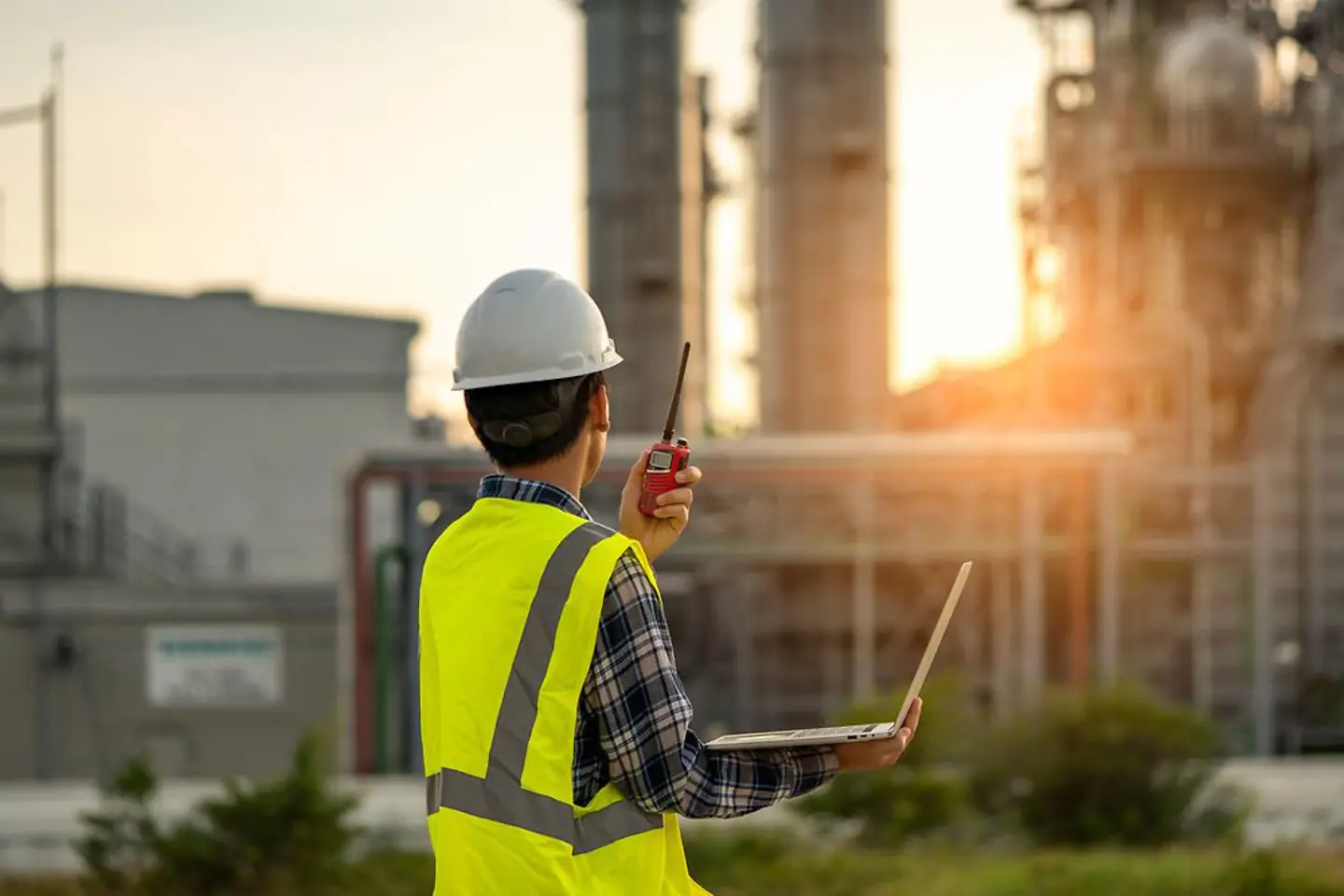 A worker in a high-visibility vest with a two-way radio and a laptop in front of the oil refinery.