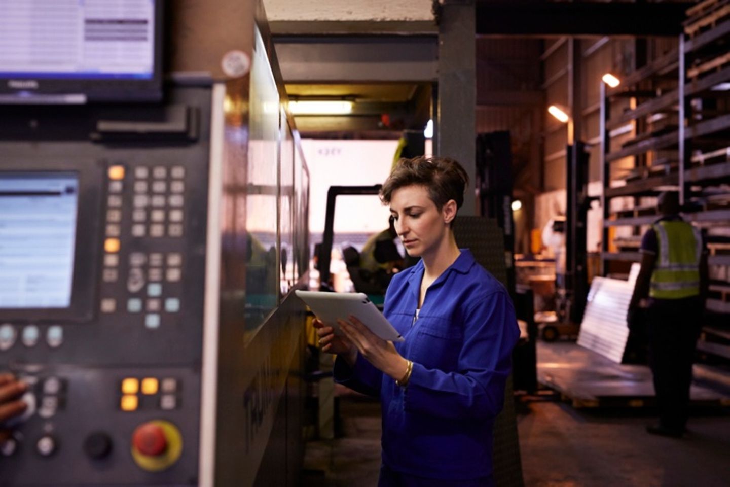 A female steel worker in a blue suit is holding a tablet in her hand.