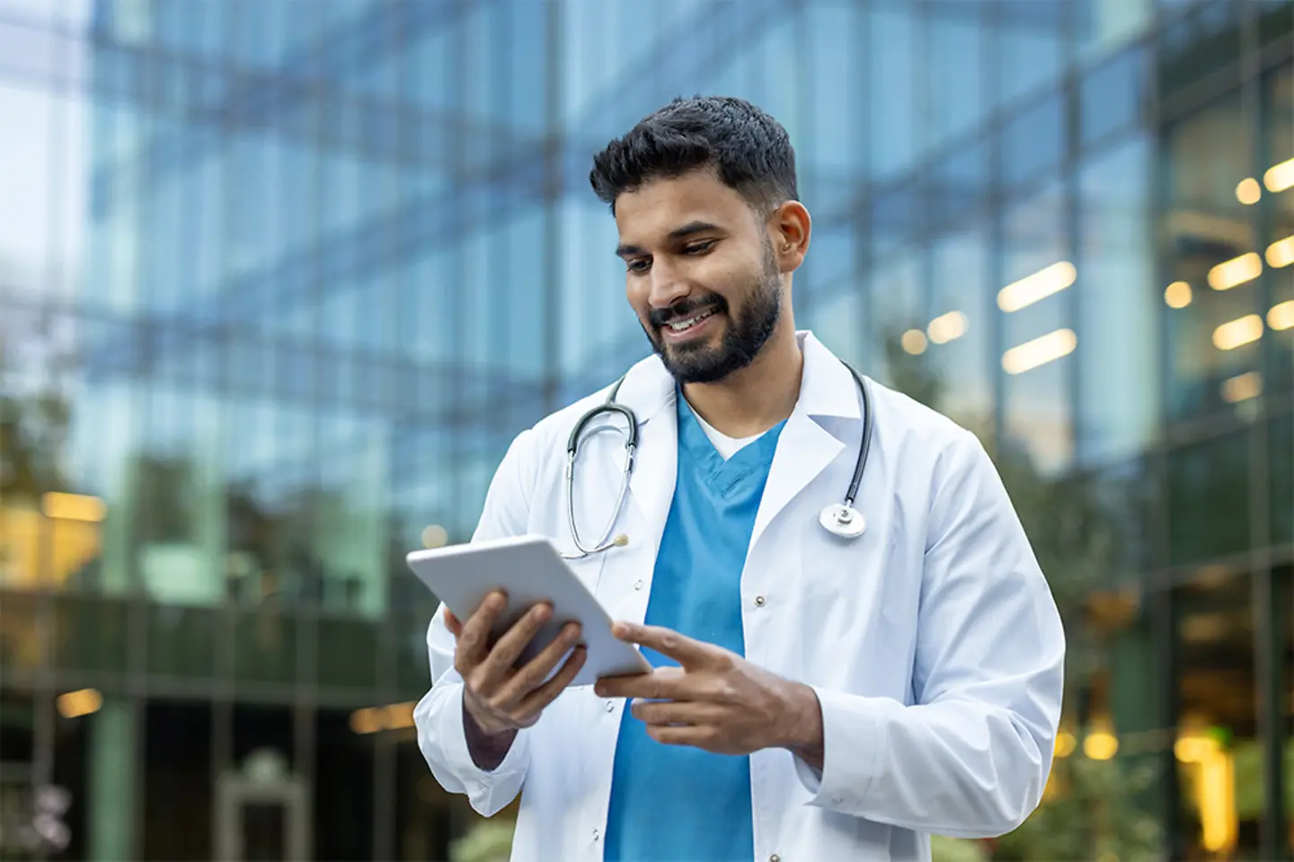 A smiling doctor uses a tablet in front of a hospital building.