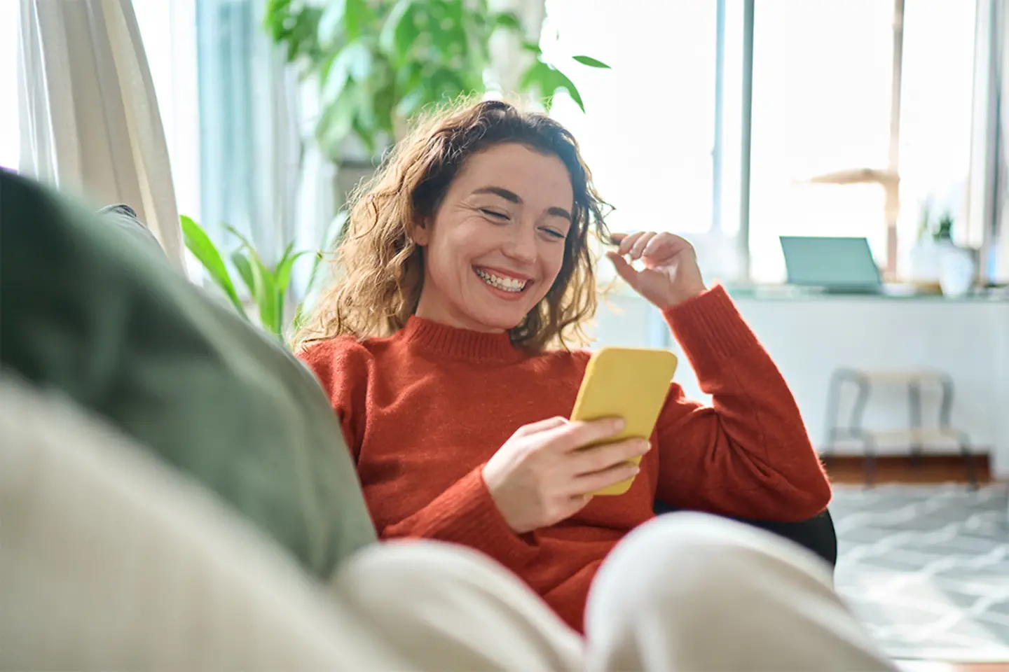 Junge Frau sitzt lächelnd auf dem Sofa und nutzt ihr Smartphone