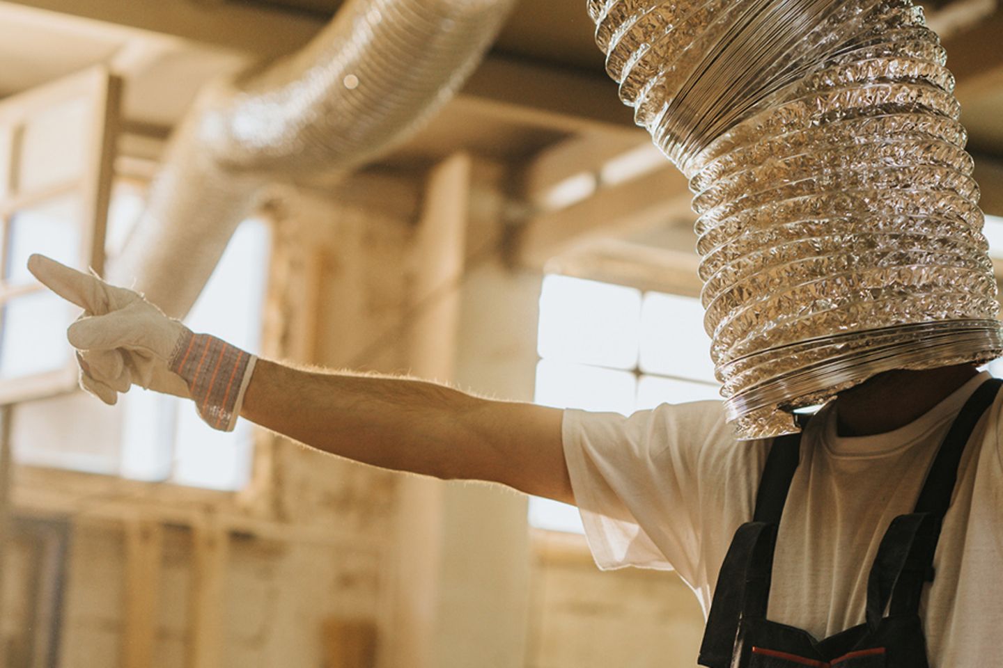 A man with working trousers, gloves and a silver pipe pulled over his head, dances quasi blind in a workshop.