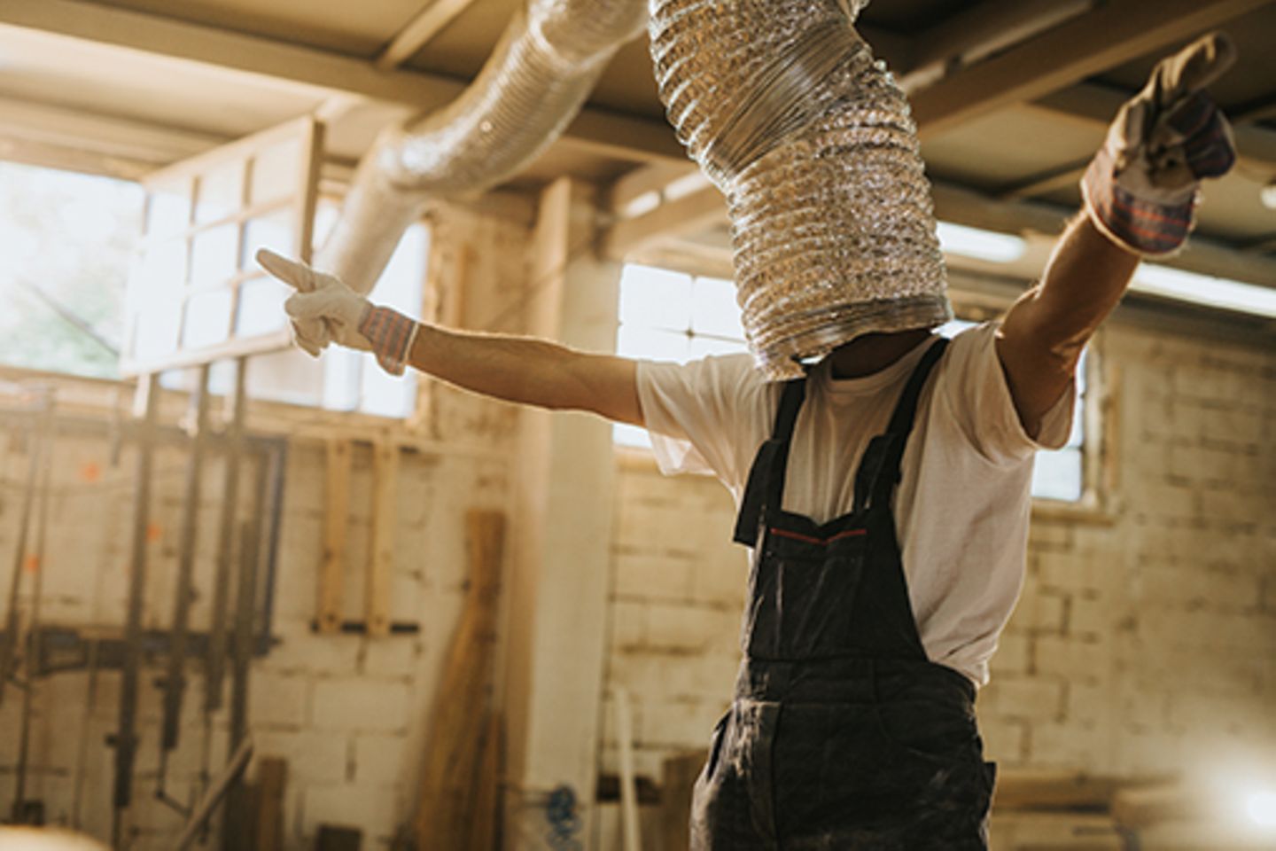 A man with working trousers, gloves and a silver pipe pulled over his head, dances quasi blind in a workshop.