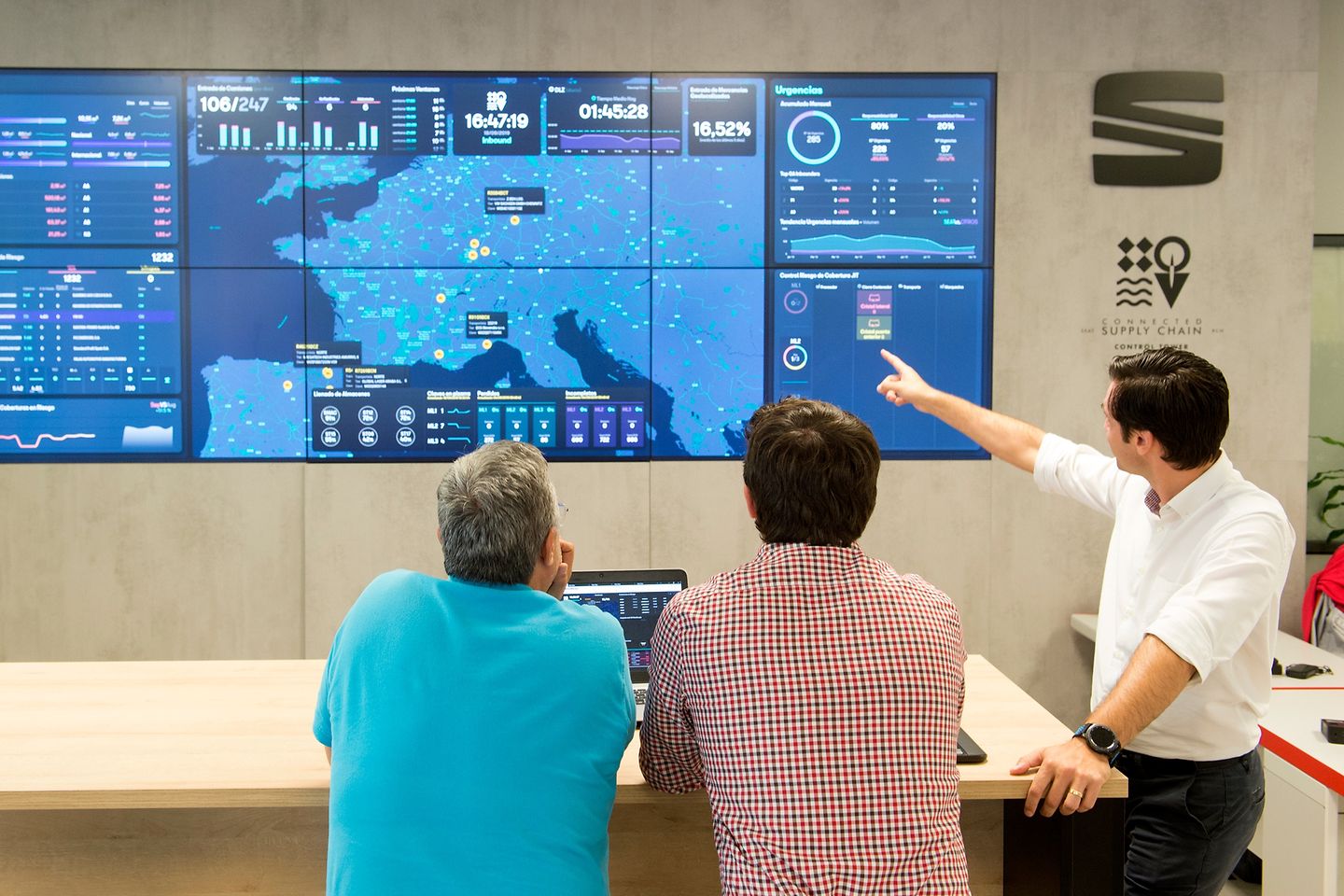 Three men look at a large dashboard and follow the current movement of goods.