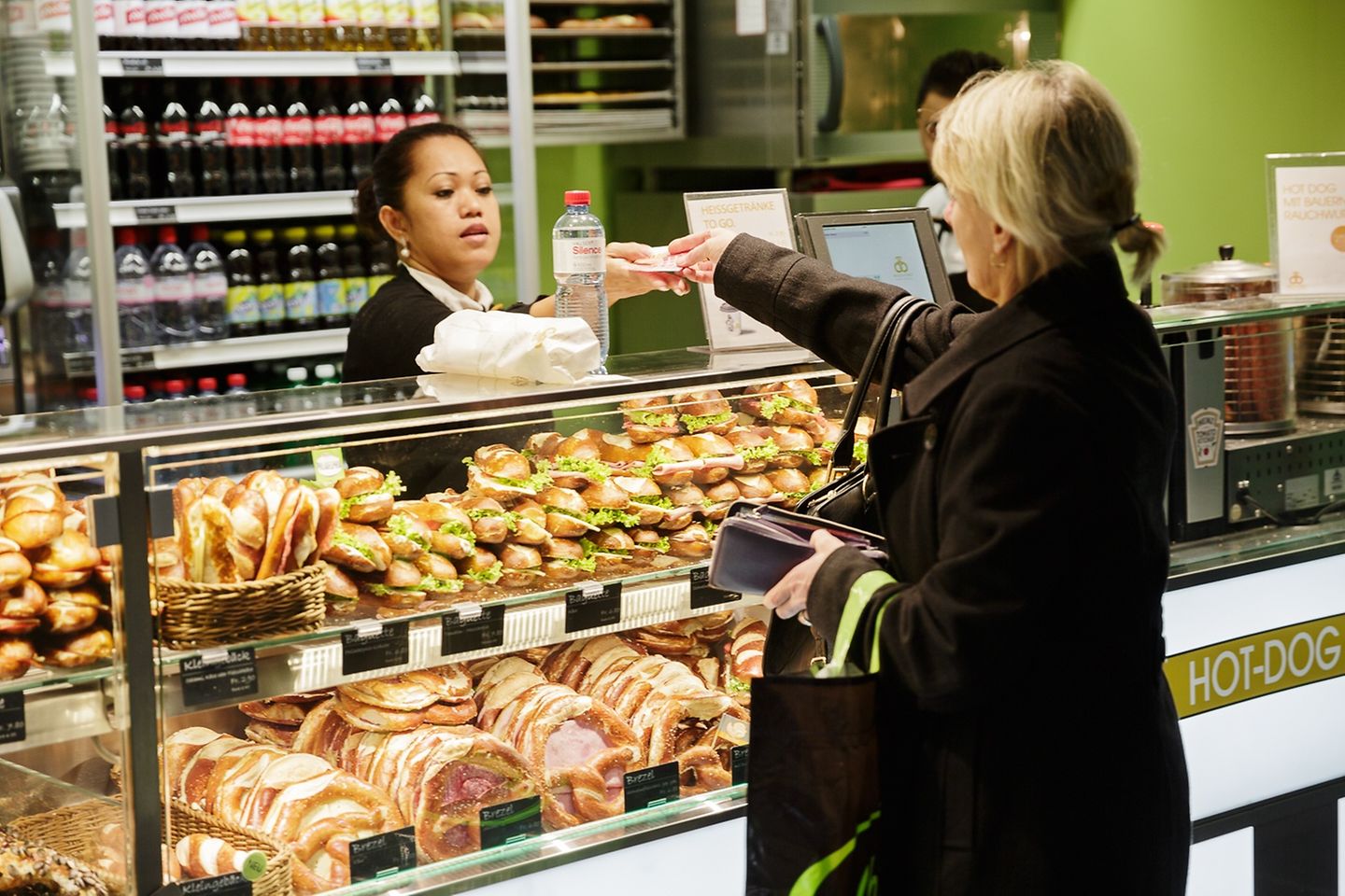 A woman in a black coat hands money to a woman behind a shop counter, which is equipped with buns.