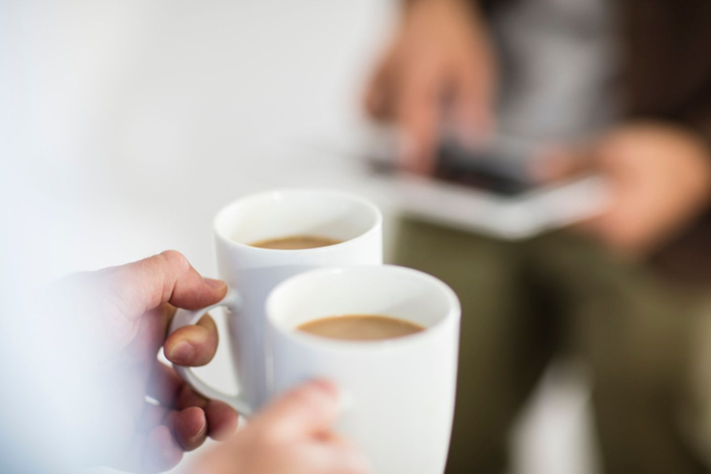 Eine Person hält zwei Kaffeetassen in den Händen, gegenüber eine andere Person mit einem Tablet in der Hand