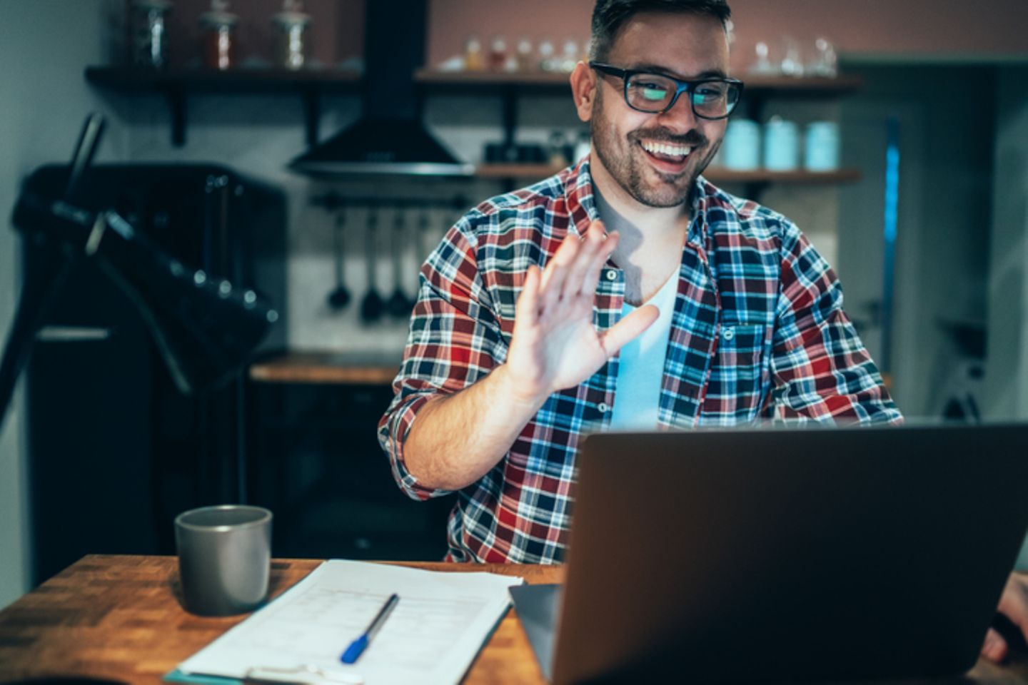 Mann sitzt am Schreibtisch und winkt jemandem am Laptop zu