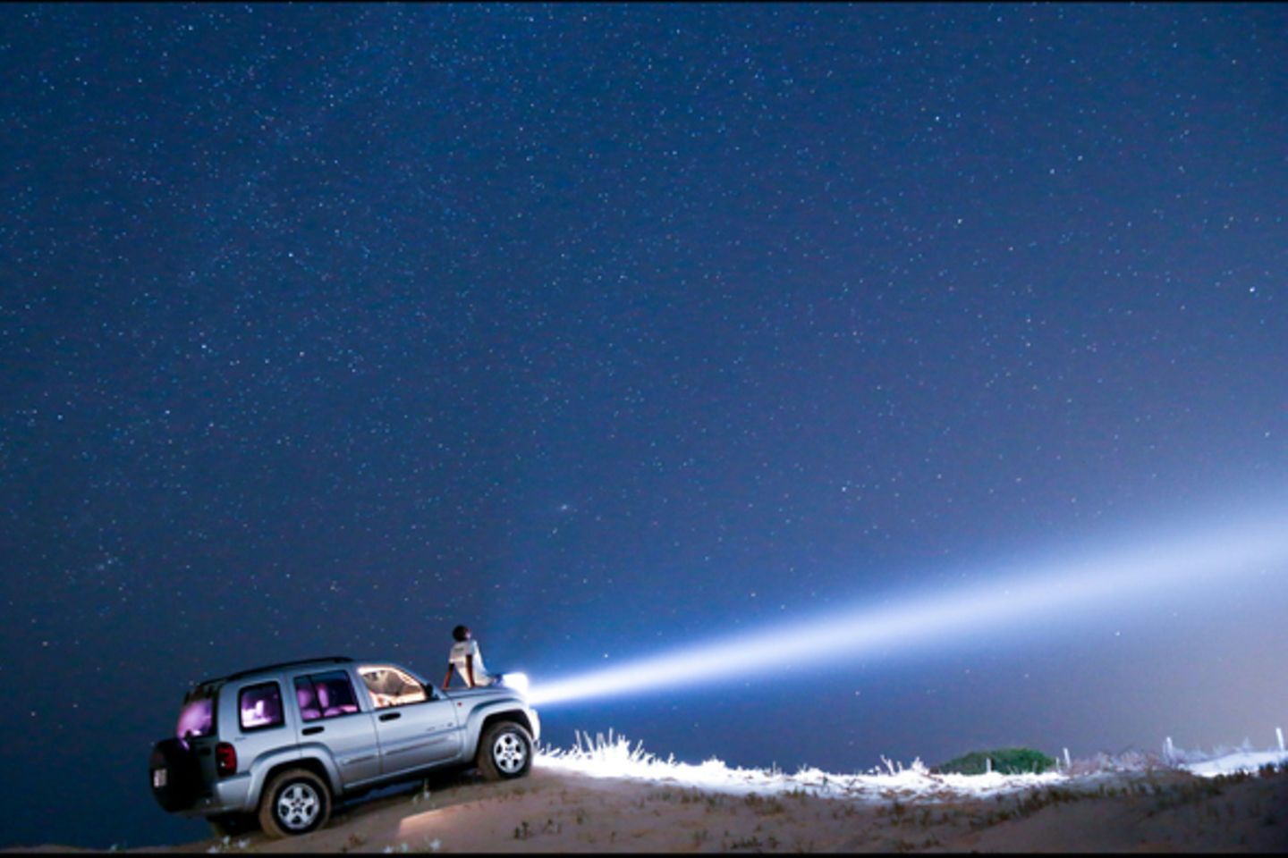 Auto steht auf einer Düne, die Scheinwerfer leuchten weit in den Nachthimmel. Eine Person sitzt auf der Motorhaube