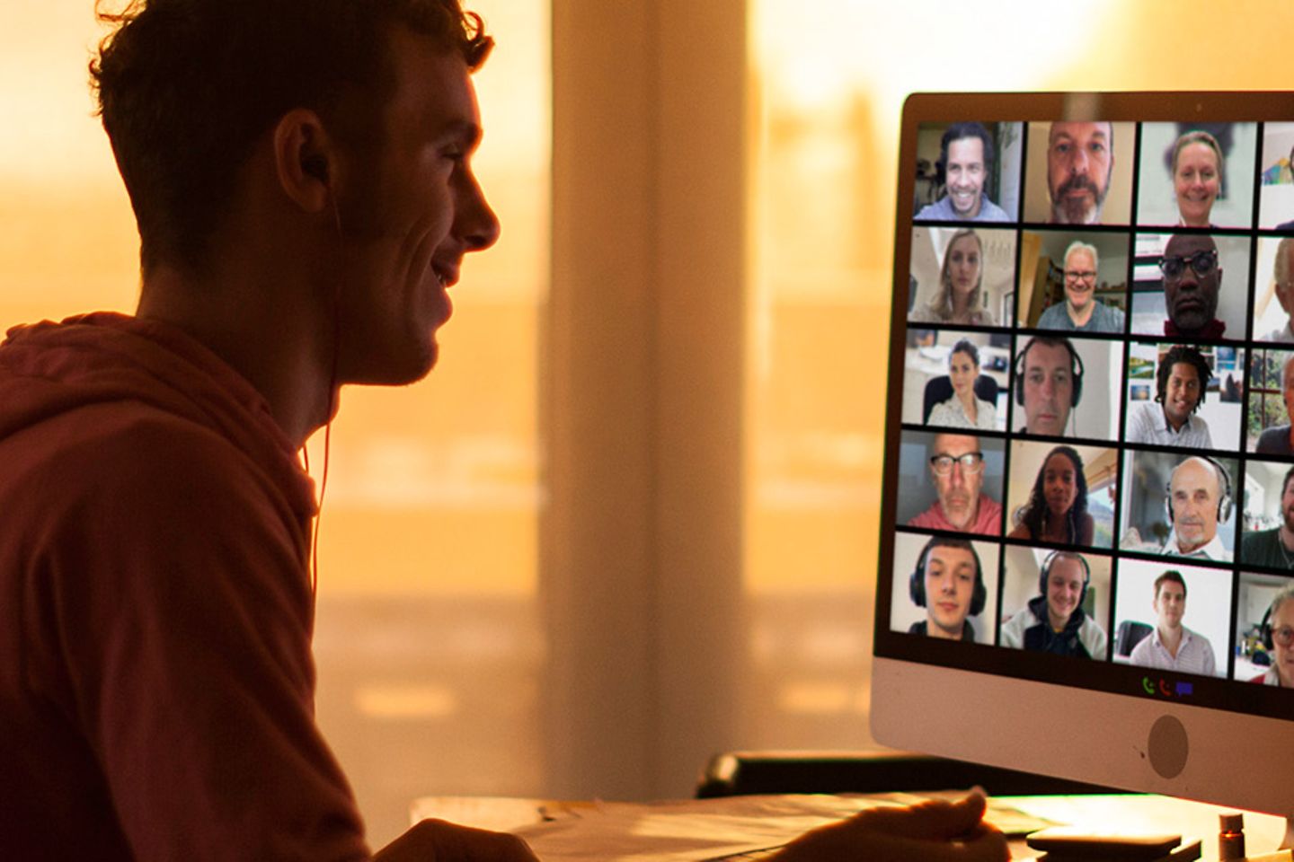 A man makes a group video call with many other people, behind him through the big windows you can see that the sun is setting