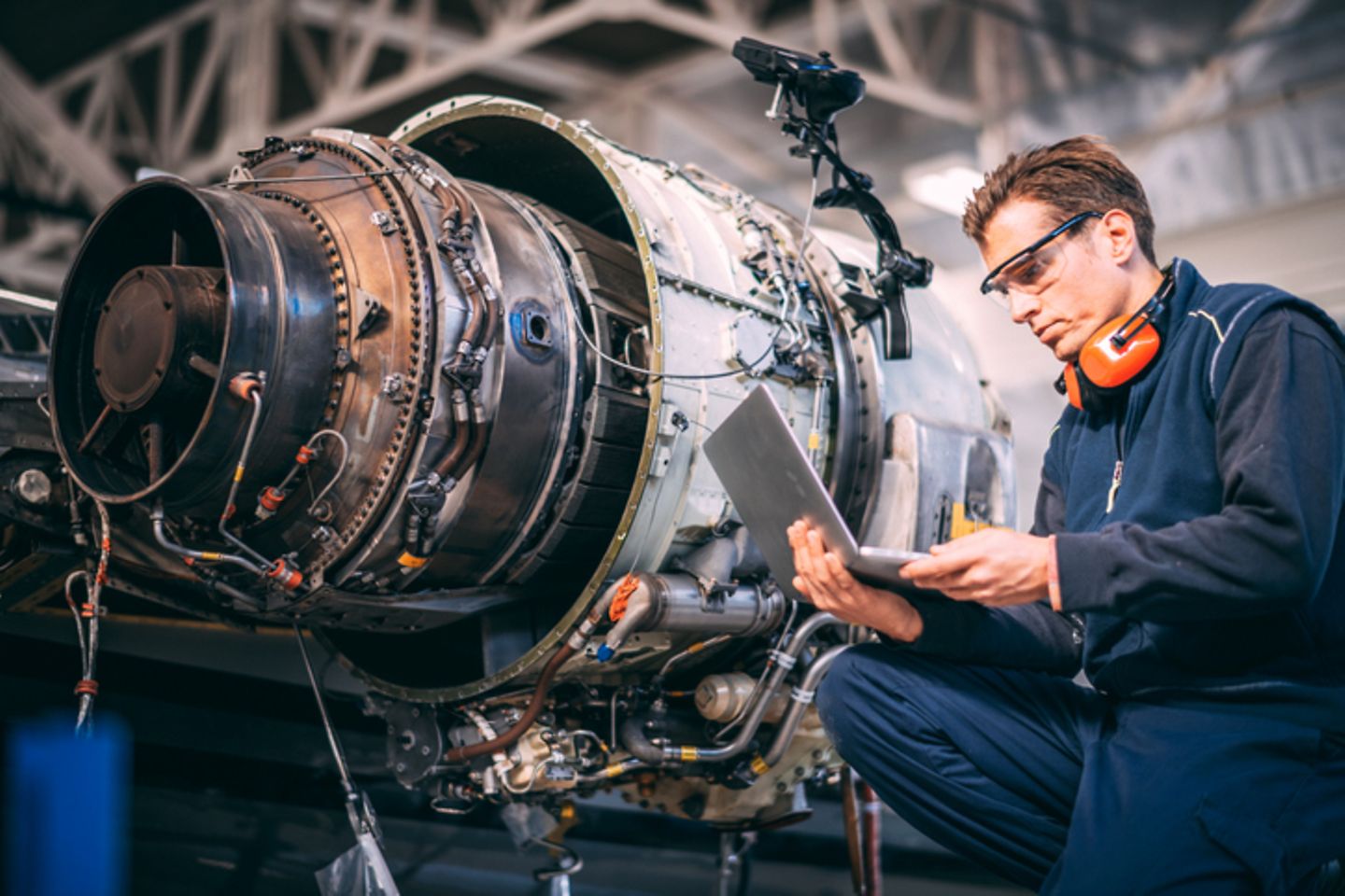 A man with a laptop in his hand kneels beside an aircraft engine.
