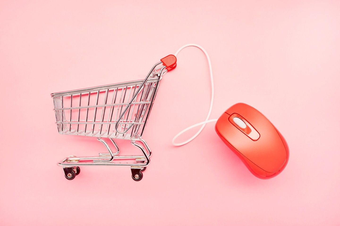 Stilleben eines kleinen Einkaufswagens und einer roten Computermaus auf rosa Hintergrund.