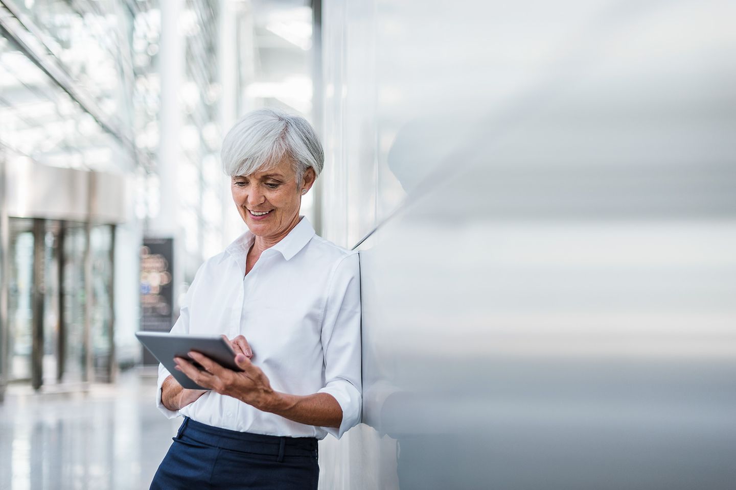 Oudere vrouw in een hal leunt tegen een zilveren muur en kijkt glimlachend op haar tablet die ze in haar hand houdt