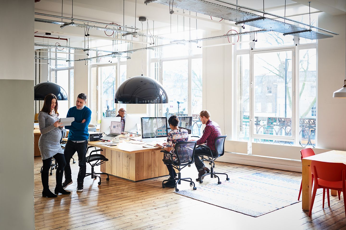 Several employees stand or sit in an office loft and look at something on a tablet or computer