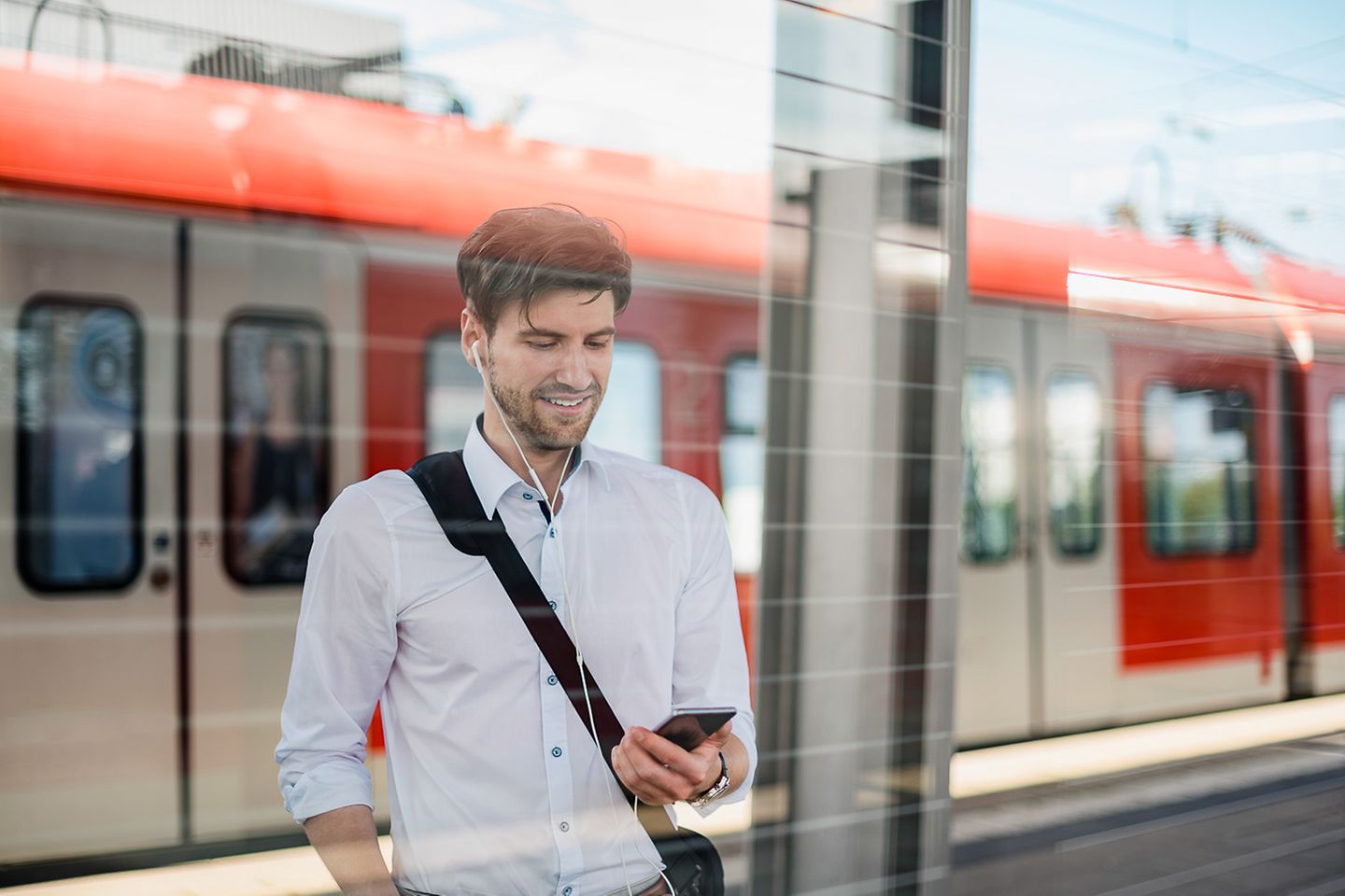 Hombre de pie en un andén mirando sonriente su smartphone. Detrás hay un tren regional.
