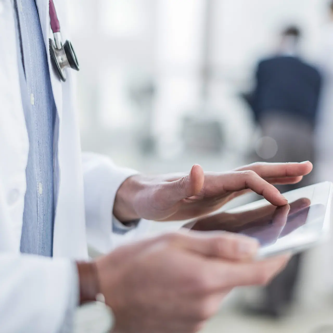 Primer plano de un médico con estetoscopio alrededor del cuello tecleando en un tablet. Hay una cara irreconocible.
