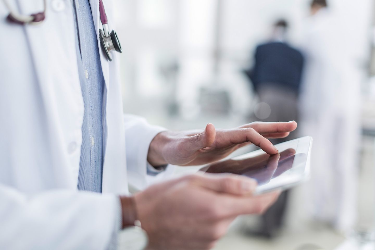 Primer plano de un médico con estetoscopio alrededor del cuello tecleando en un tablet. Hay una cara irreconocible.