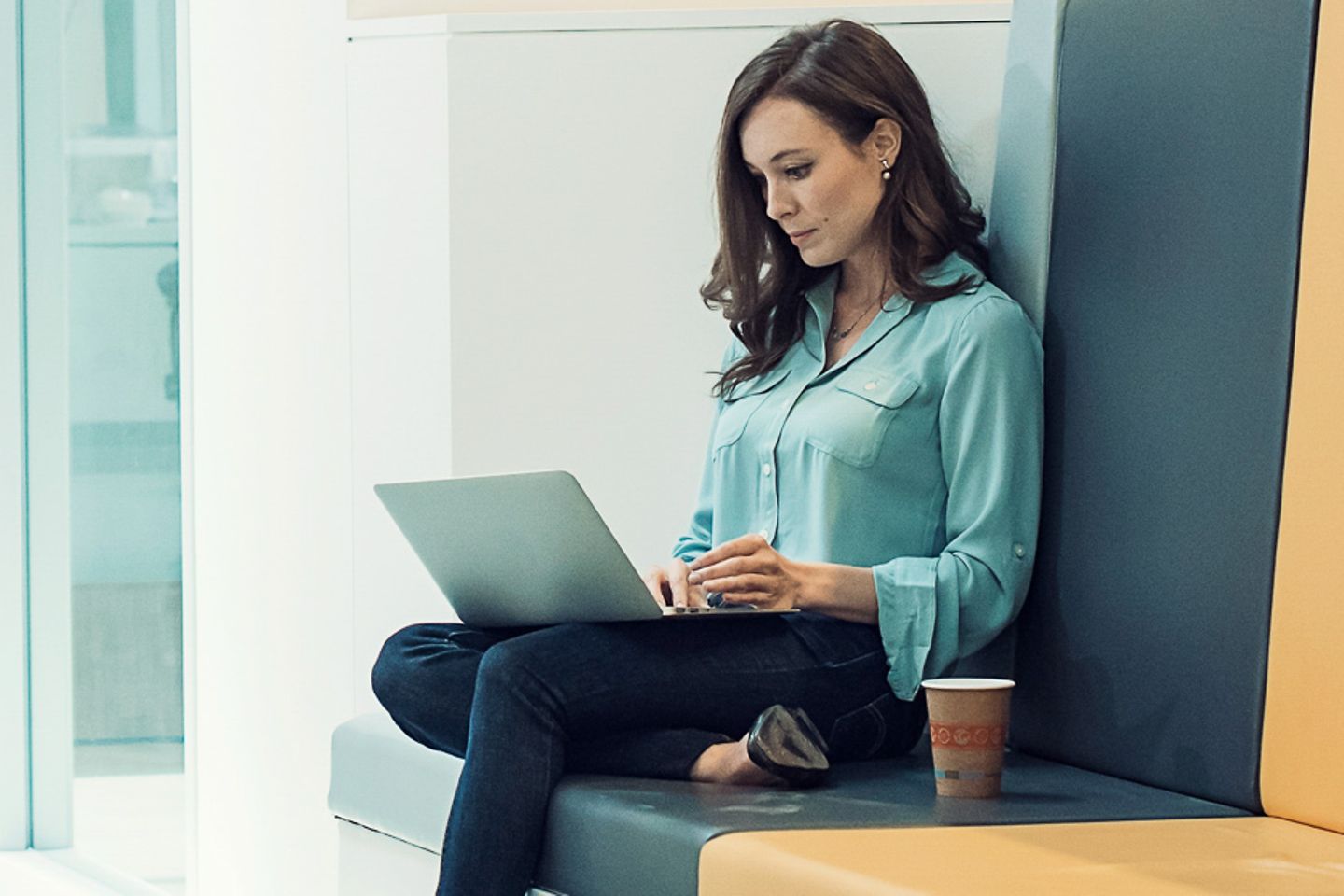 一位年轻女士坐在明亮的办公室走廊里，膝上放着一台笔记本电脑