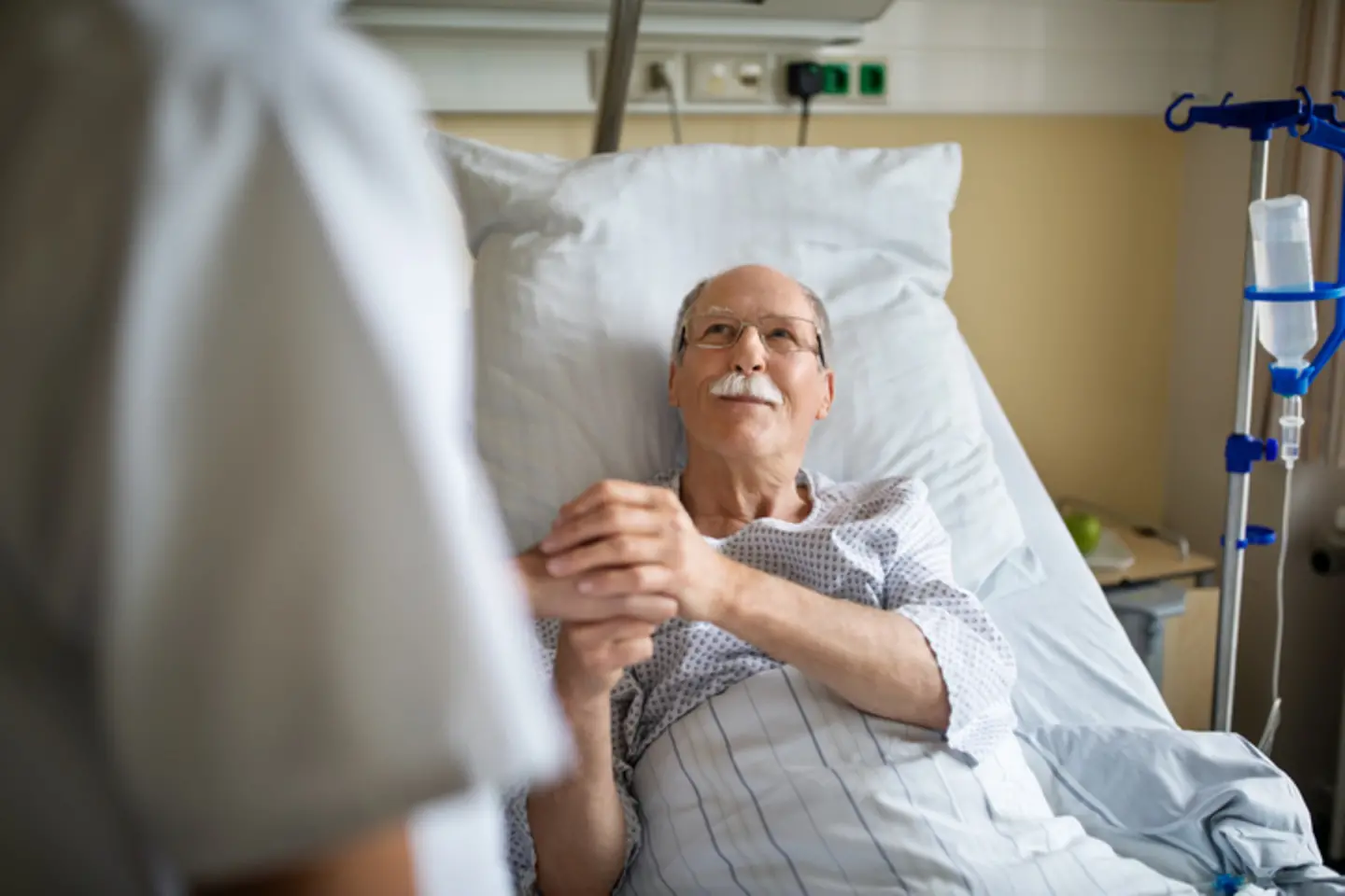 An elderly man lies on a hospital bed and holds a nurse’s hand