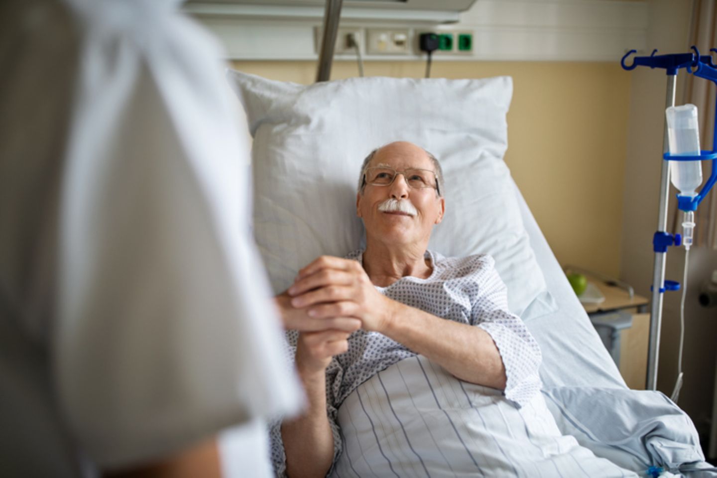 An elderly man lies on a hospital bed and holds a nurse’s hand