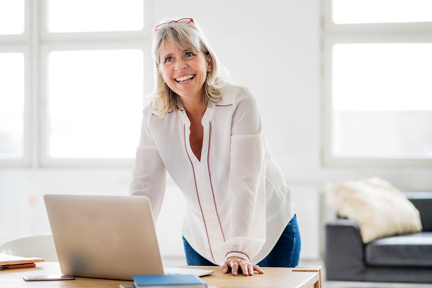 Uma mulher de meia idade está sorrindo e recostada em uma mesa de escritório, na qual se encontra um laptop aberto