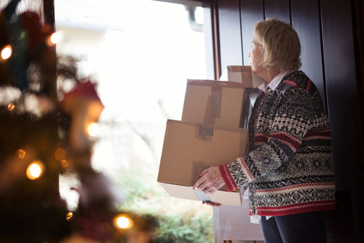 Older woman on a front door holding two packages, in the foreground is a christmas tree