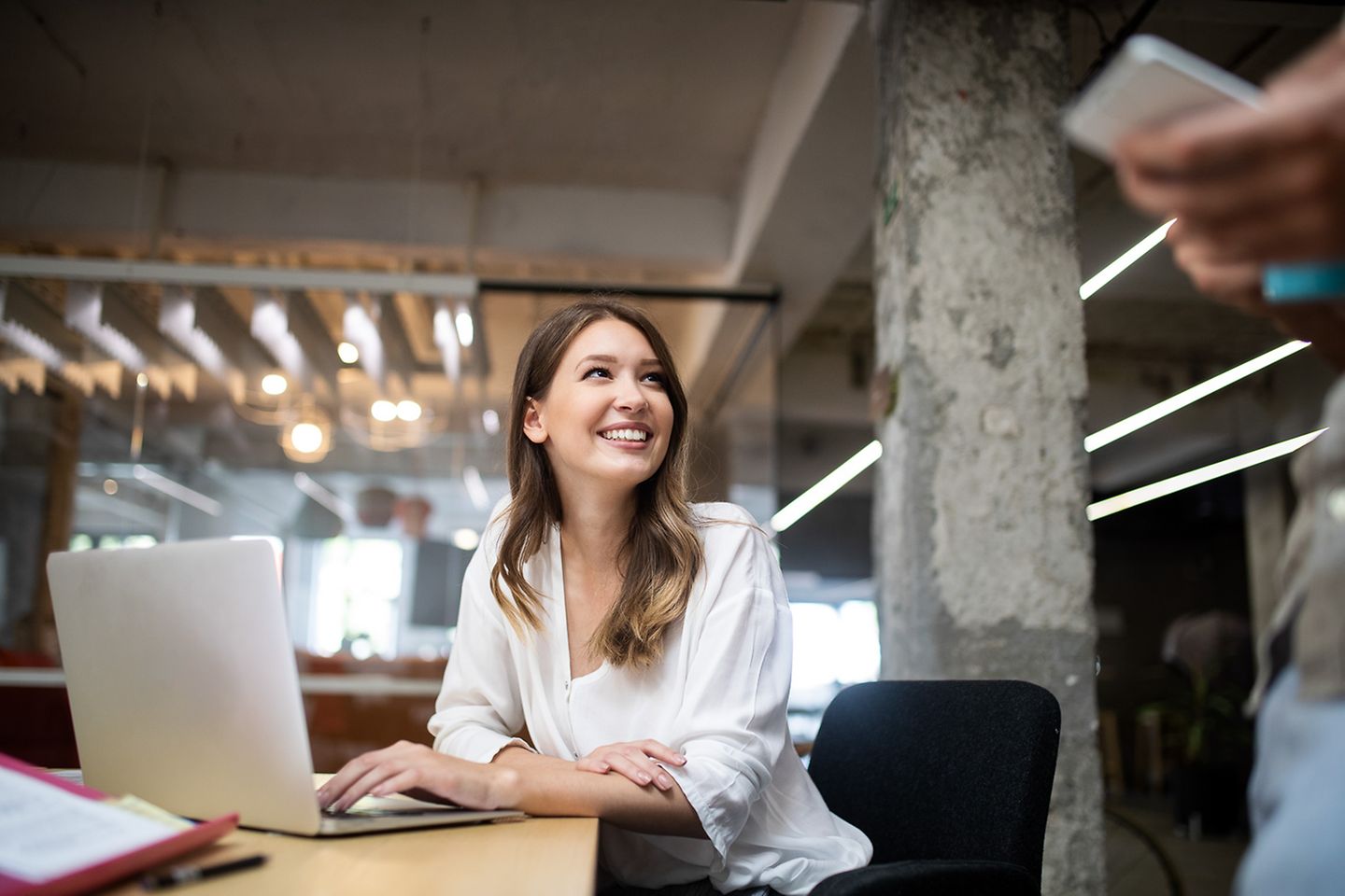 Mulher sorridente sentada num escritório usando um laptop e olhando para um colega de pé