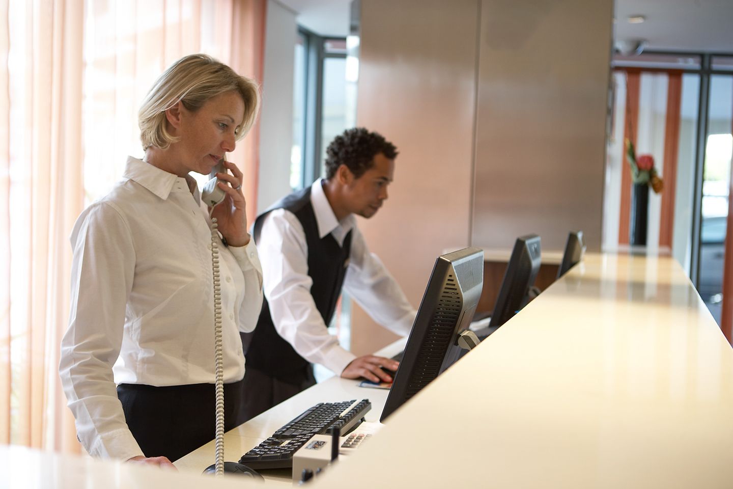 Uma recepcionista faz uma ligação atrás da recepção do hotel. Seu colega verifica os dados no computador