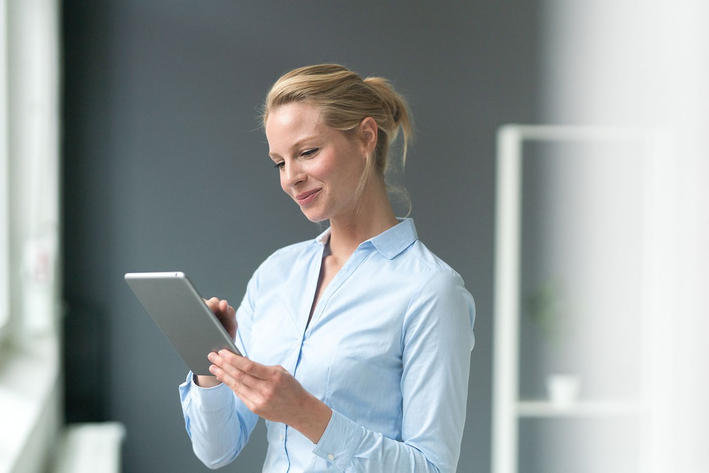 Eine Frau in einem blauen Hemd blickt lächelnd auf ein Tablet.