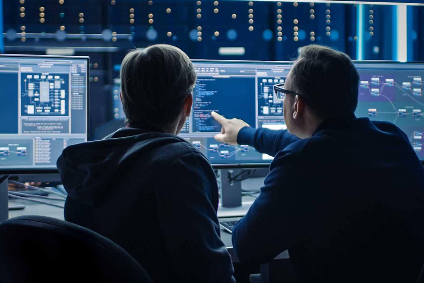 Two men in front of two large monitors discussing data