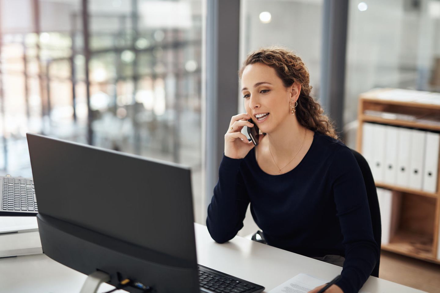 Mulher jovem sentada em seu laptop e falando ao telefone.