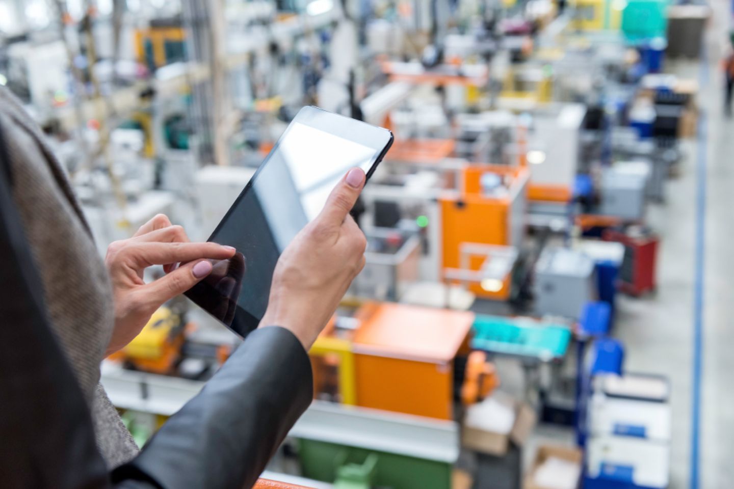 Mains qui pianotent sur une tablette. En arrière-plan, les contours d’un hall d’usine avec des machines.