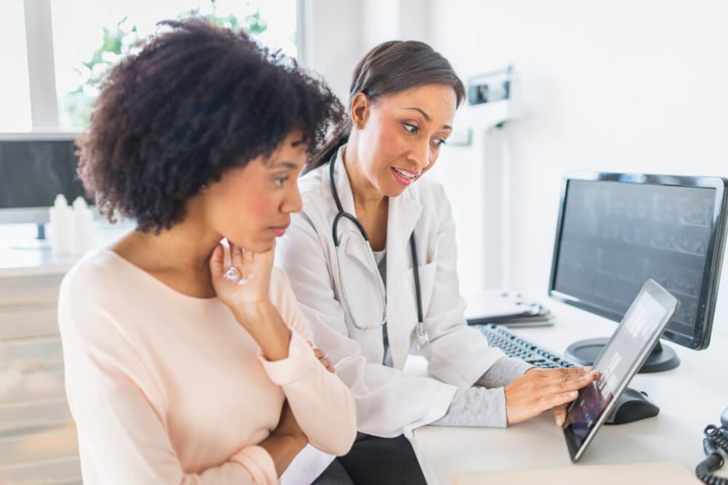 Two women, one of which is a doctor are sitting at a desk and are looking at an iPad 