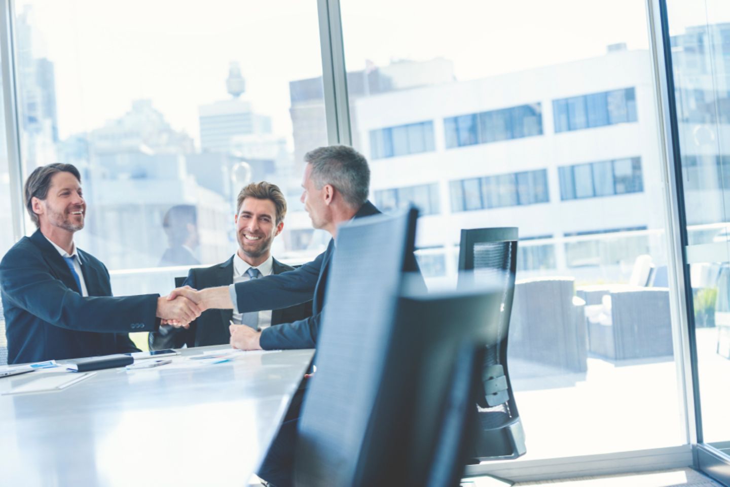 Drei männer sitzen an einem Bürotisch und lachen, zwei schütteln sich die Hände