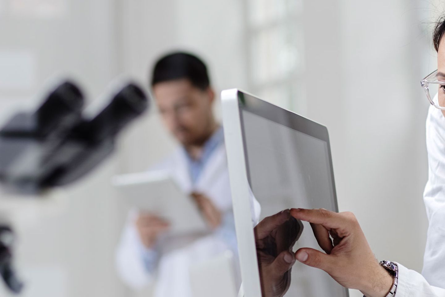 Three people in a laboratory are wearing white gowns. Blurry image of a microscope can be made out.