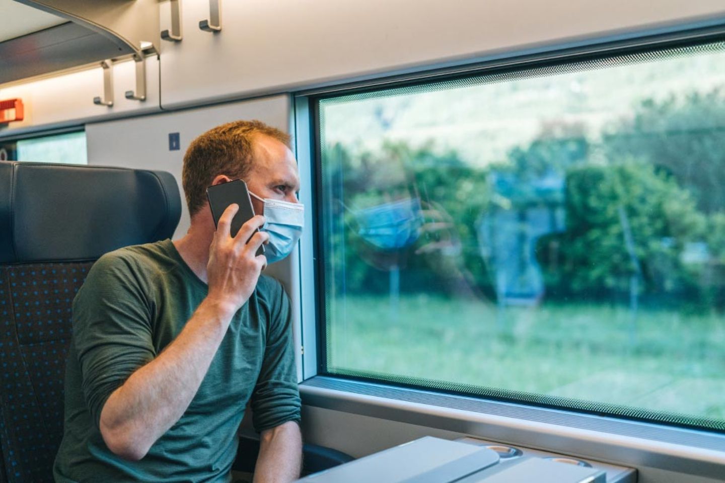 Mann sitzt im Zug mit einer Maske auf dem Telefon und schaut aus dem Fenster