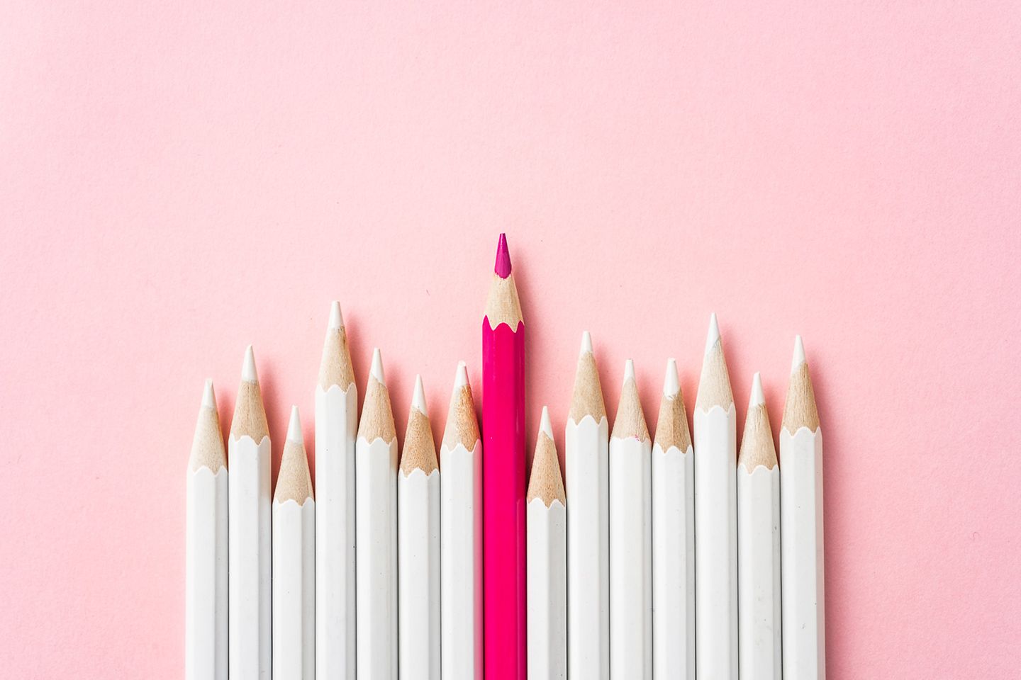 Reihe weißer Buntstifte mit einem pinkfarbenen Buntstift in der Mitte vor rosa Hintergrund.