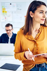 Geschäftsfrau steht in modernem Büro mit Tablet und Stift in den Händen, ihr Kollege sitzt im Hintergrund am Laptop