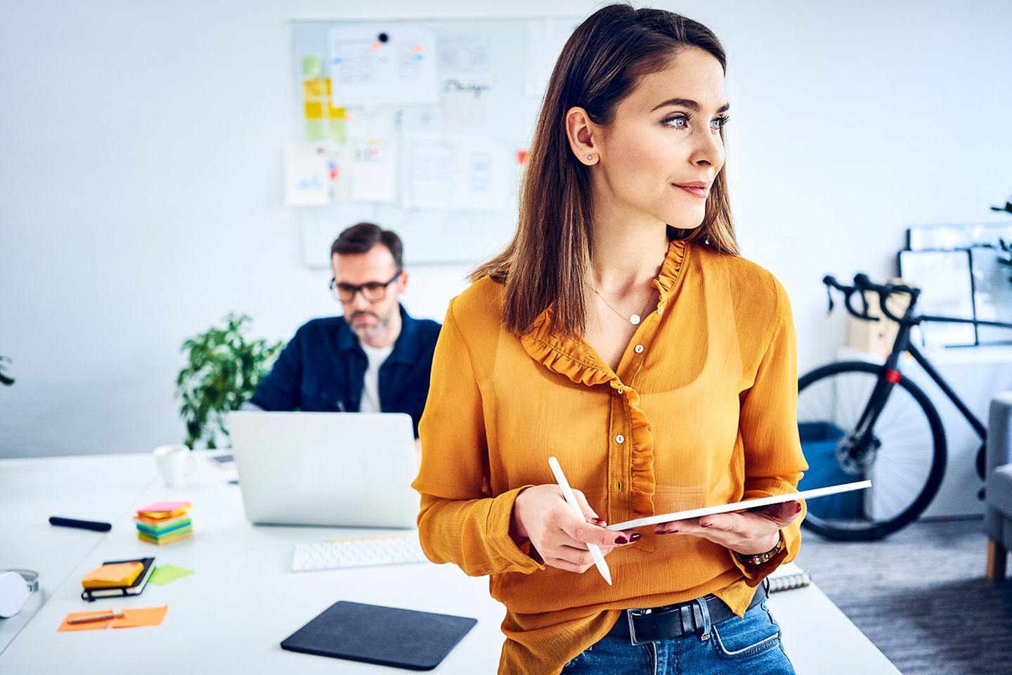 Geschäftsfrau steht in modernem Büro mit Tablet und Stift in den Händen, ihr Kollege sitzt im Hintergrund am Laptop