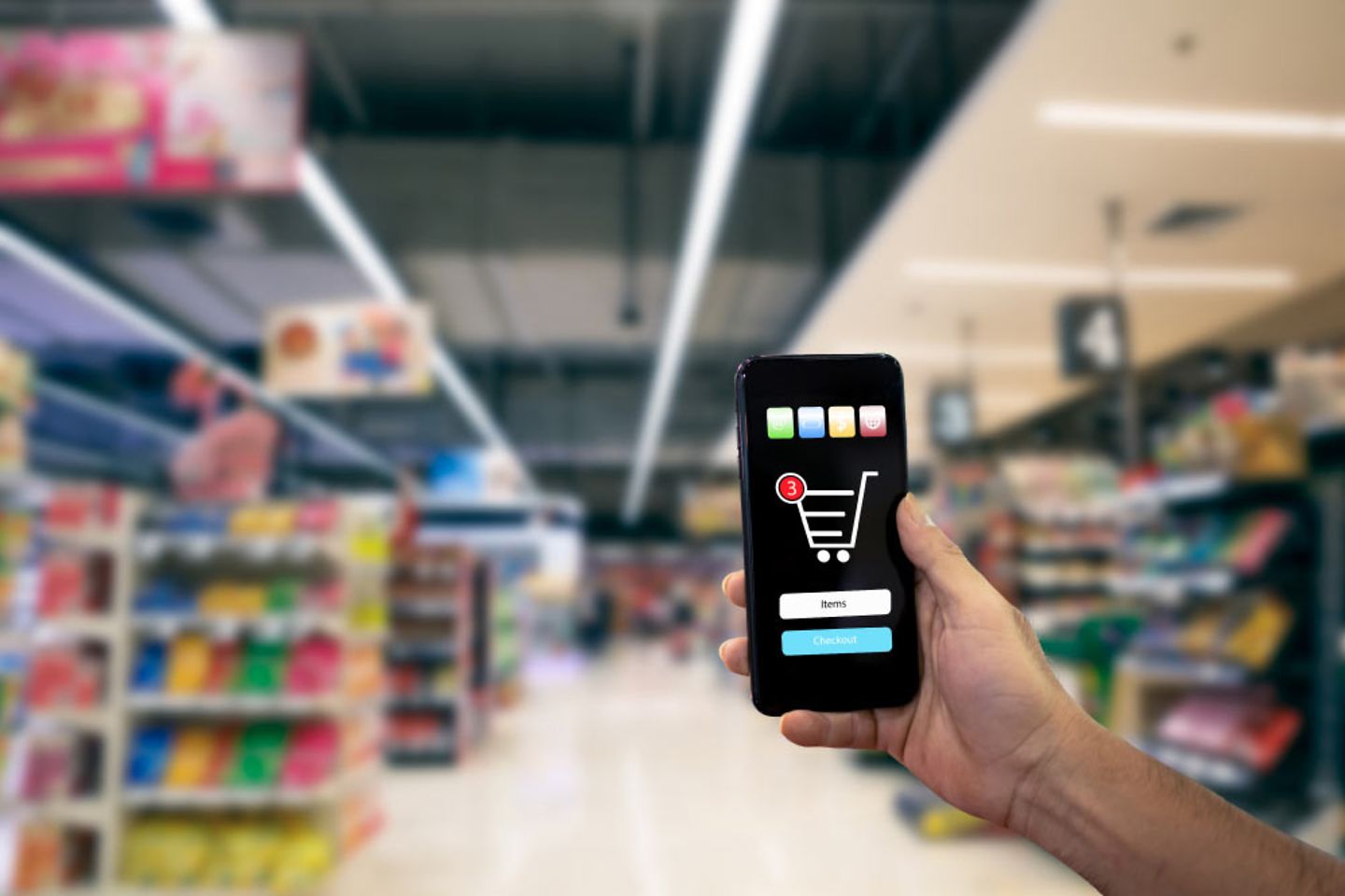 One hand holds a smartphone showing a shopping cart, in the background are supermarket shelves