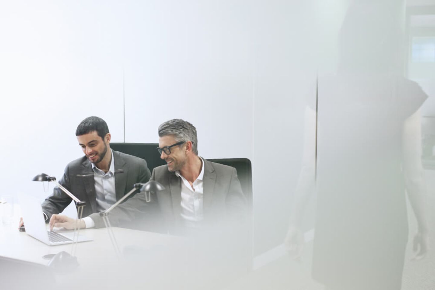 Two men in suits sit at a desk and smile