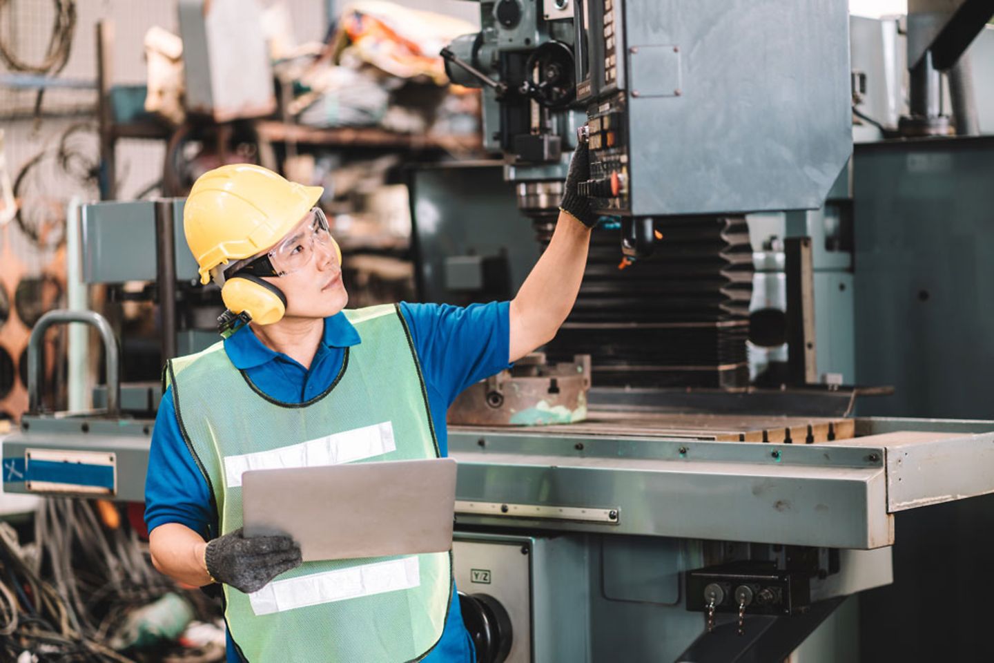 Worker in safety work wear with yellow helmet and ear muff using laptop in factory