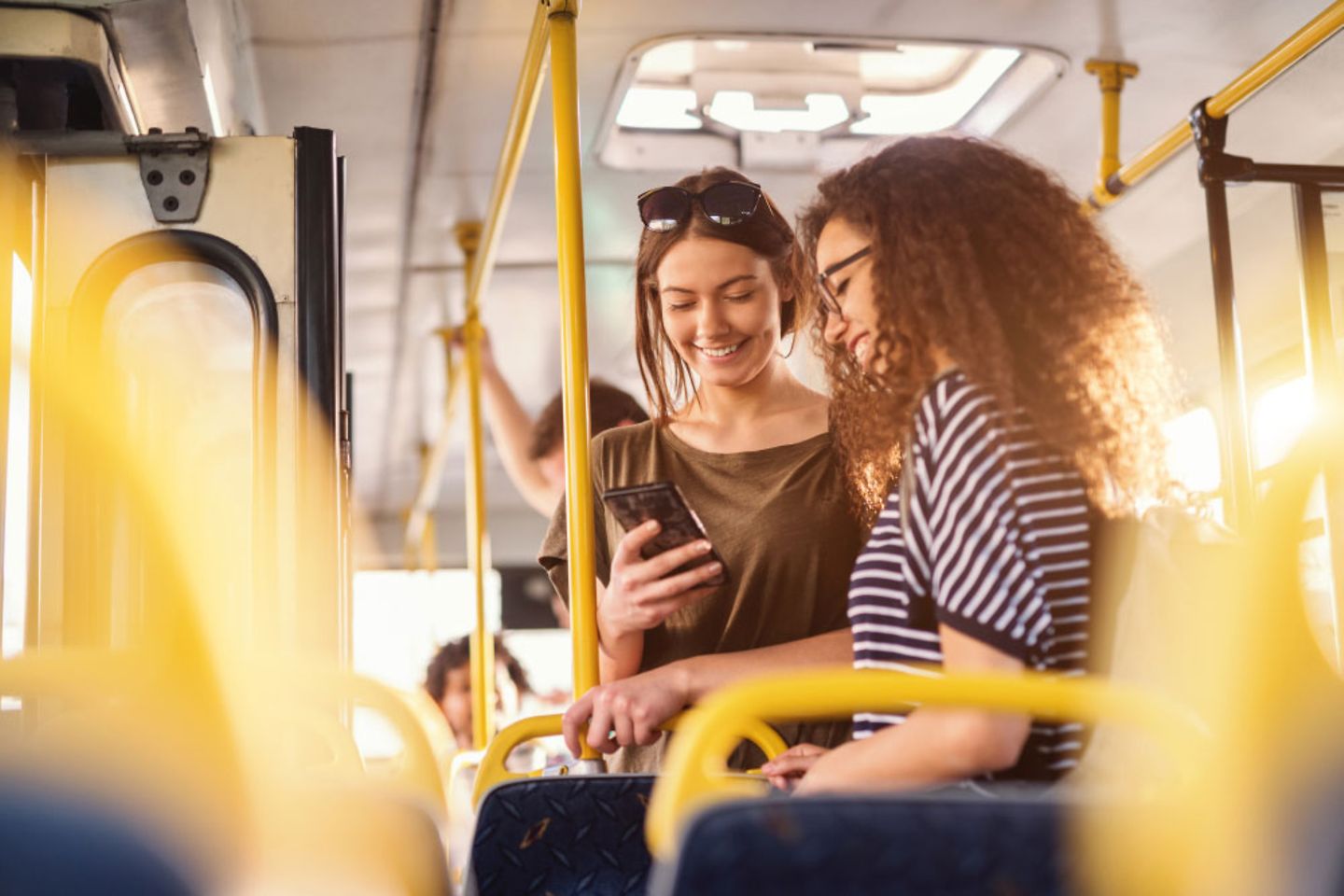 Zwei Mädchen stehen in einem öfftentlichen Verkehrsmittel und schauen auf ein Smartphone