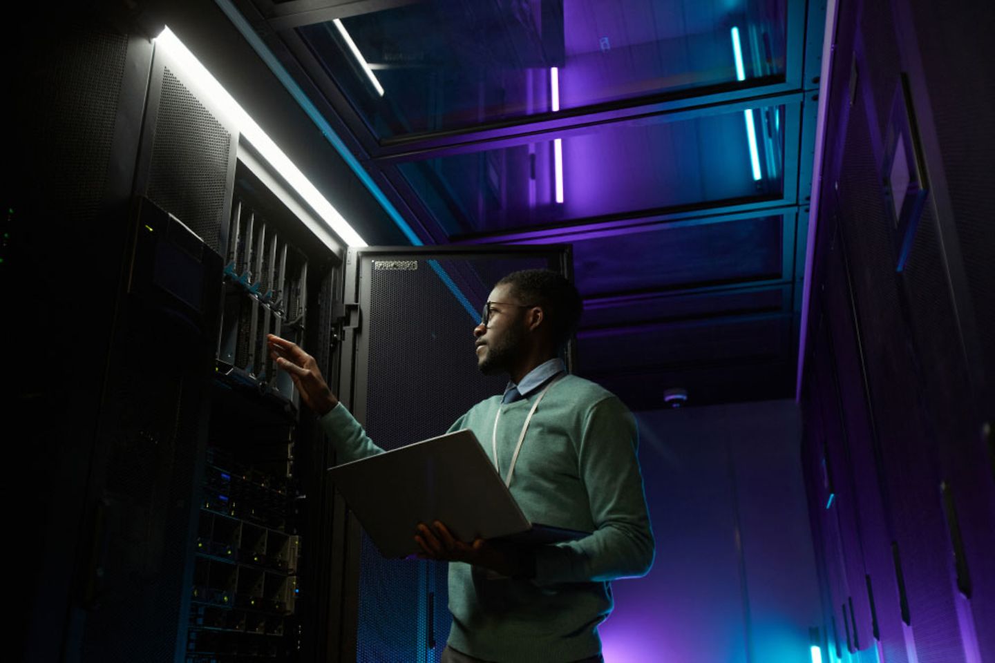 Um homem está parado com um laptop em uma sala de servidor escura em um gabinete aberto