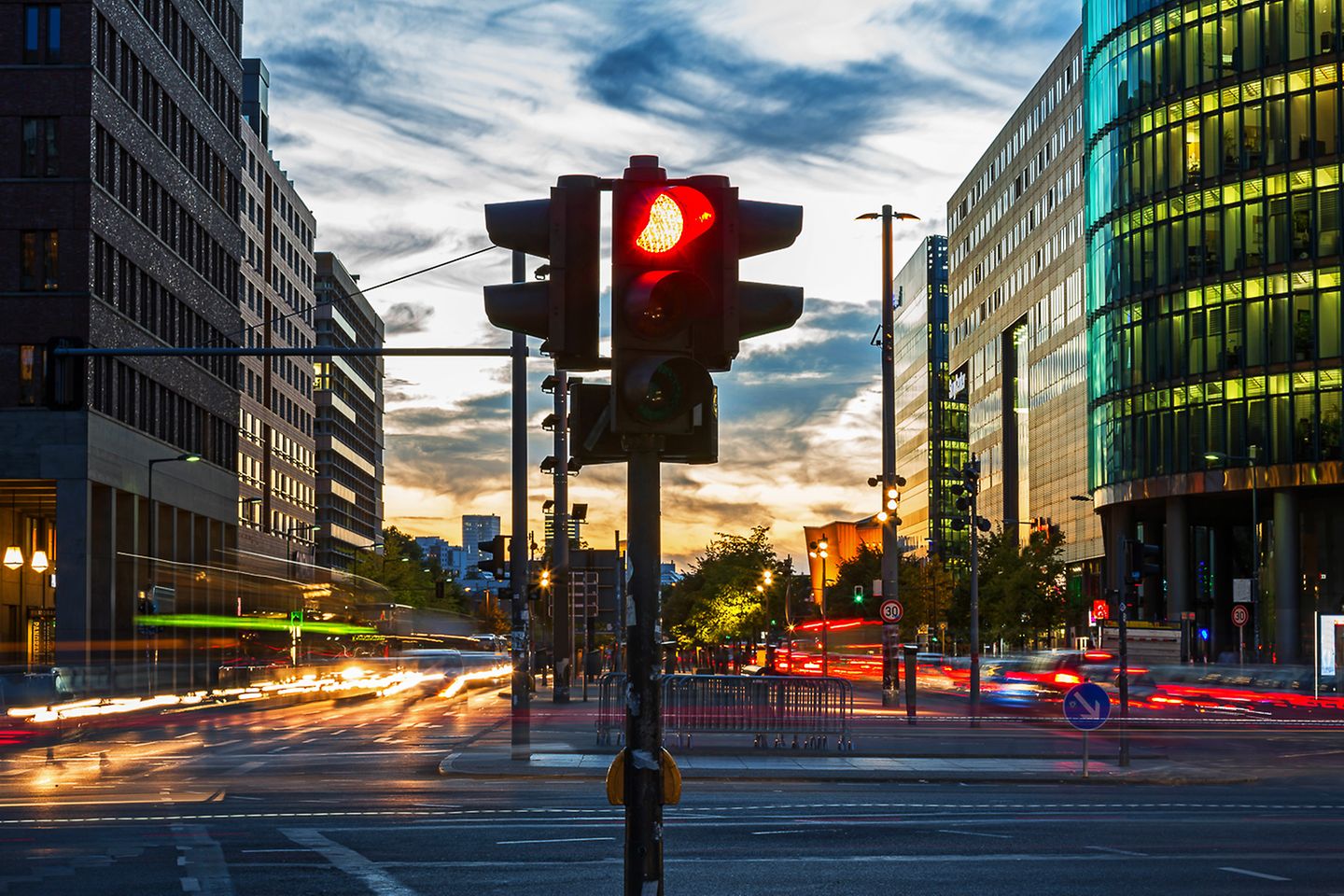 Befahrene Straßenkreuzung in der Dämmerung. In der Bildmitte steht eine rote Ampel.