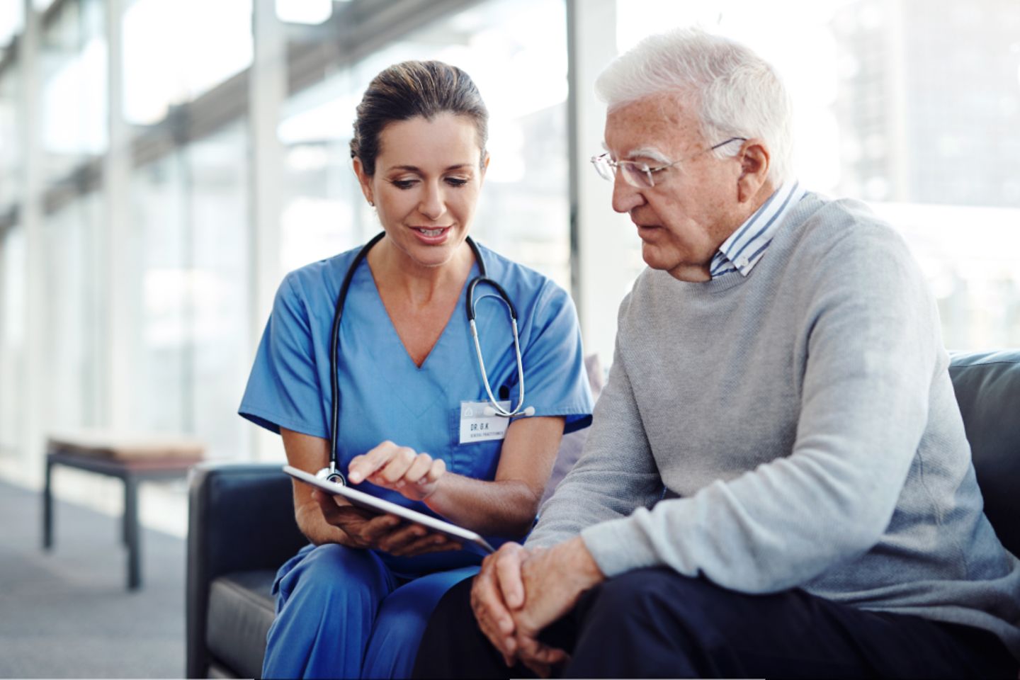 Empleada del hospital con bata azul muestra algo en una tableta a un hombre de pelo canoso