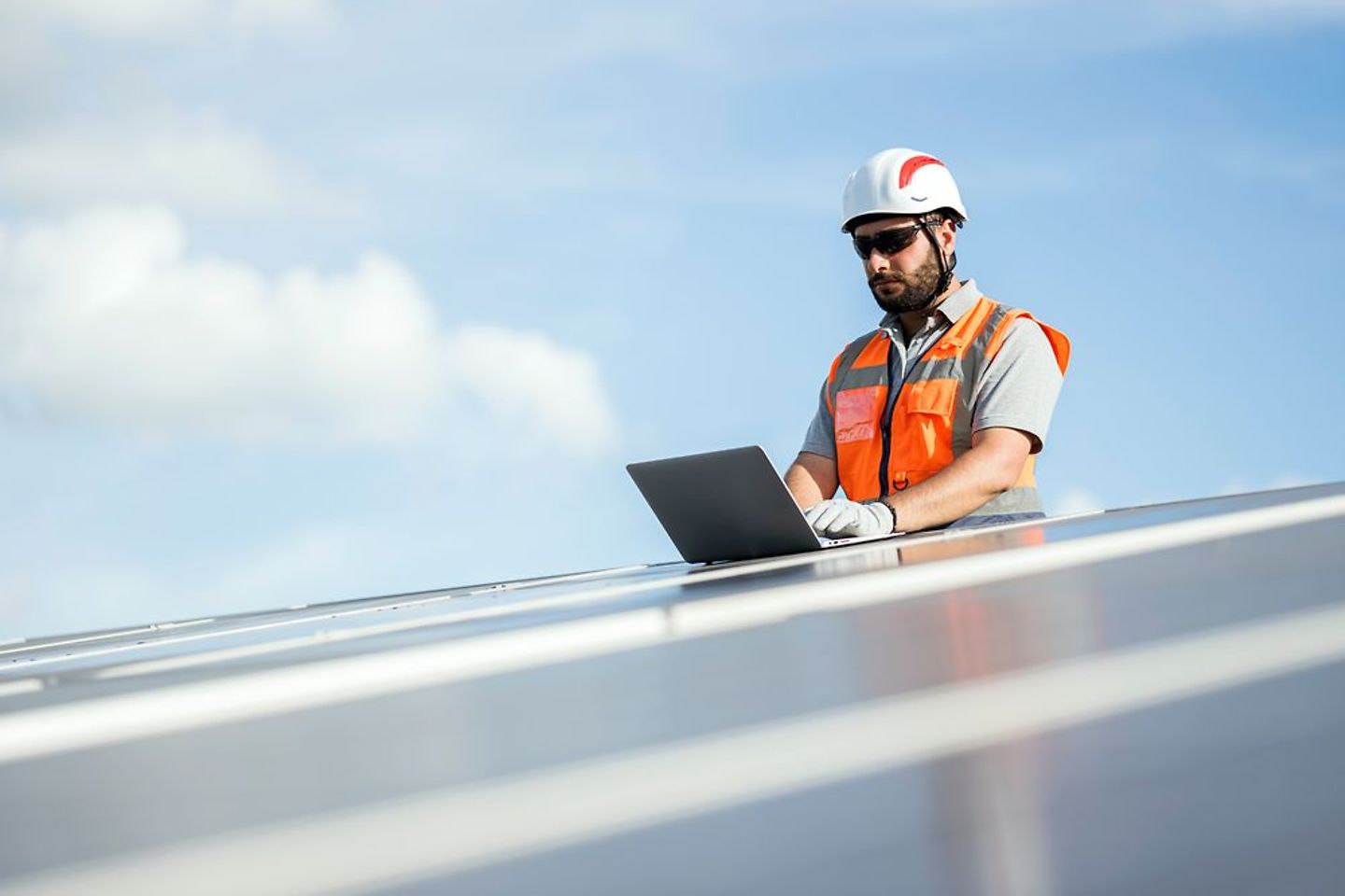 A male solar service engineer use a laptop while standing on a ladder next to a roof