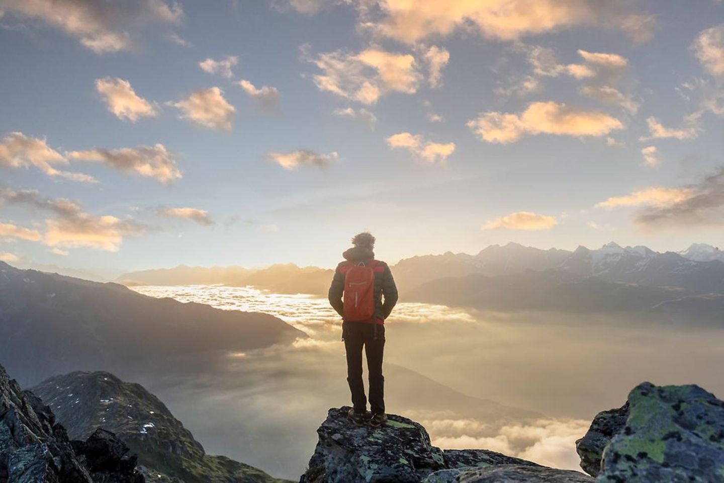 Mann mit Rucksack steht auf einem Berg vor der aufgehenden Sonne
