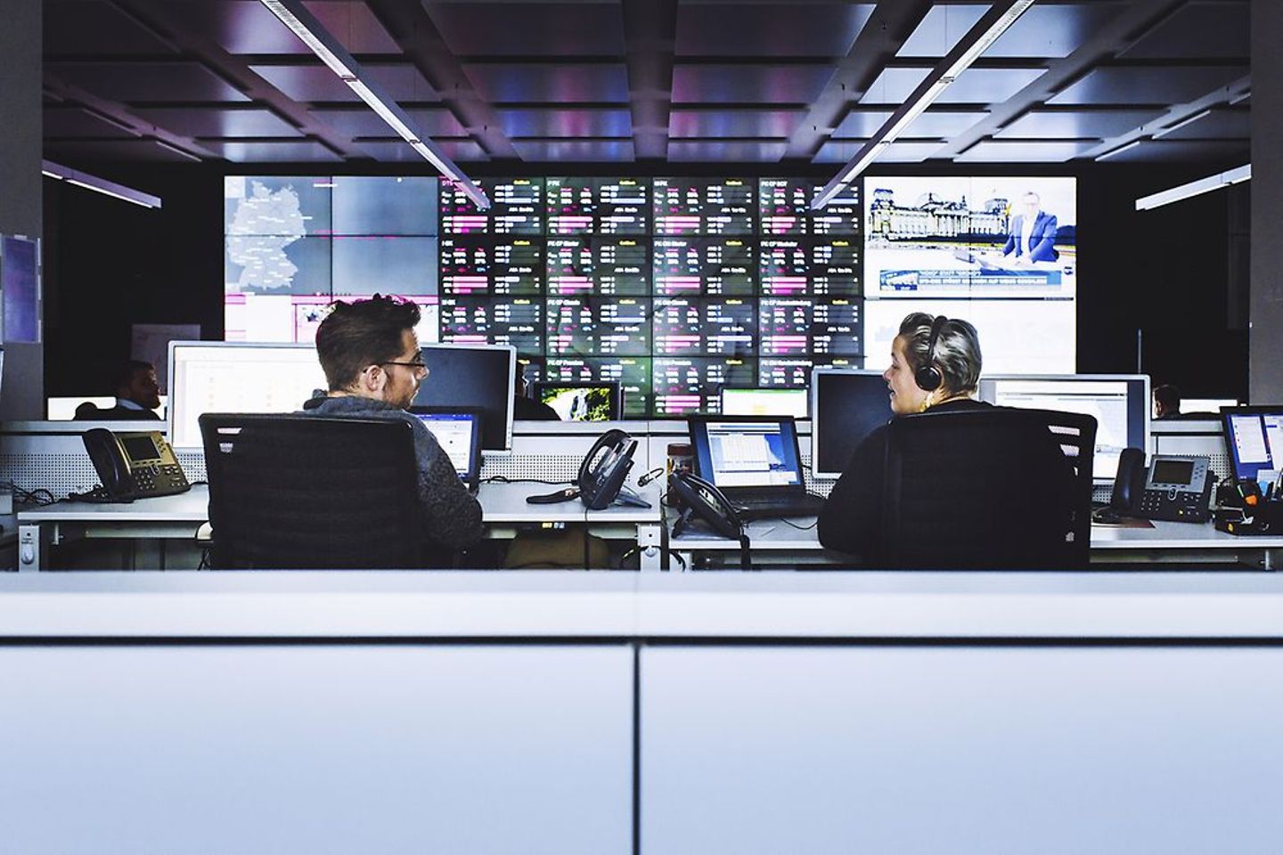 Two people sitting in a surveillance room in front of many monitors