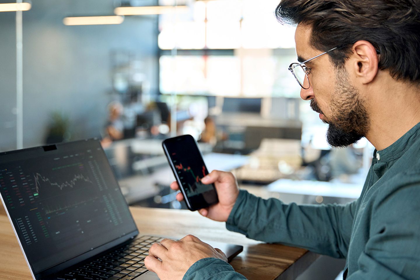 Investment Broker vor dem Laptop im Büro mit Mobile Phone analysiert Finanzkennzahlen