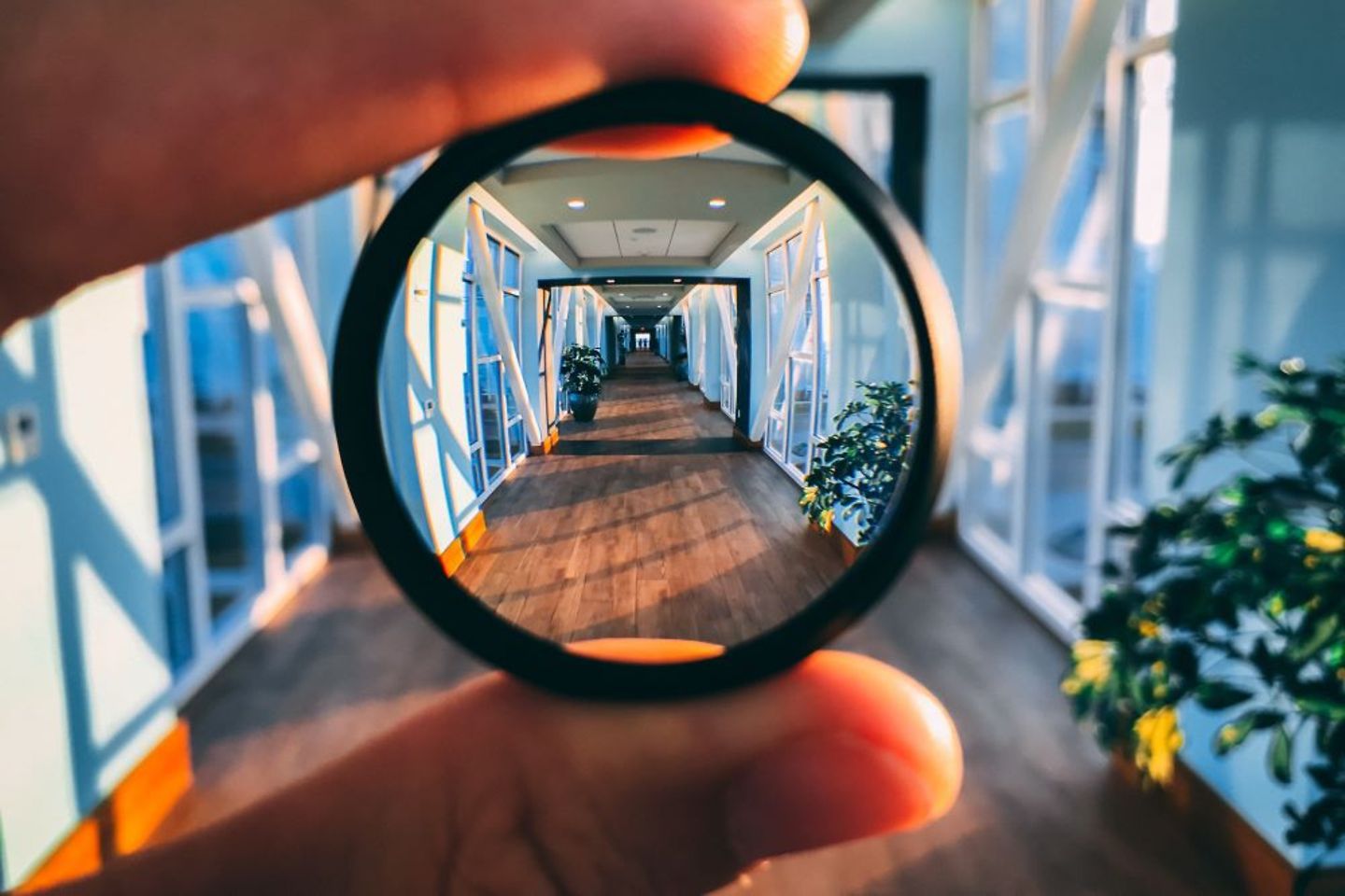 Zwei Finger halten ein Objektiv mit einem Zoom auf eine gläserne Brücke in einem Gebäude.
